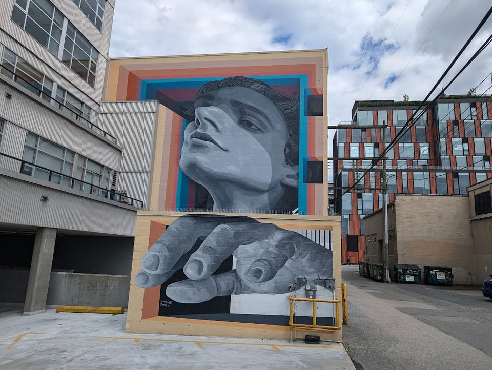 A cluster of apartment buildings and squat industrial buildings. At the center of it all is a grayscale mural on two blocks, one taller than the other. The upper block shows a person's face looking up and to the right. The lower block shows a hand reaching out. The overall effect is of a 3-D representation of a person reaching out.