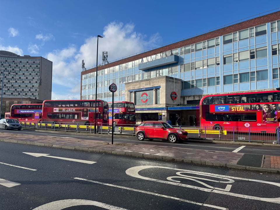 The exterior of Morden Underground station, taken from across the road. The station is square and boxy, with a glass facade and a large Tube roundel. There are several red buses and a red car on the road between me and the station.