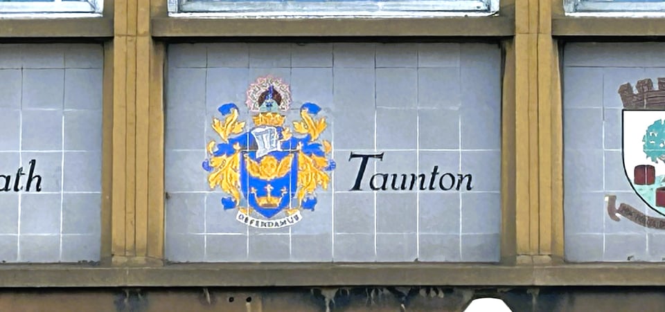 Close up on the tiled panel for Taunton. The tiles are grey and square, forming a horizontal rectangle. The coat of arms for Taunton, featuring a knight's helm, a cherub and a crown, is painted on, next to the word Taunton.