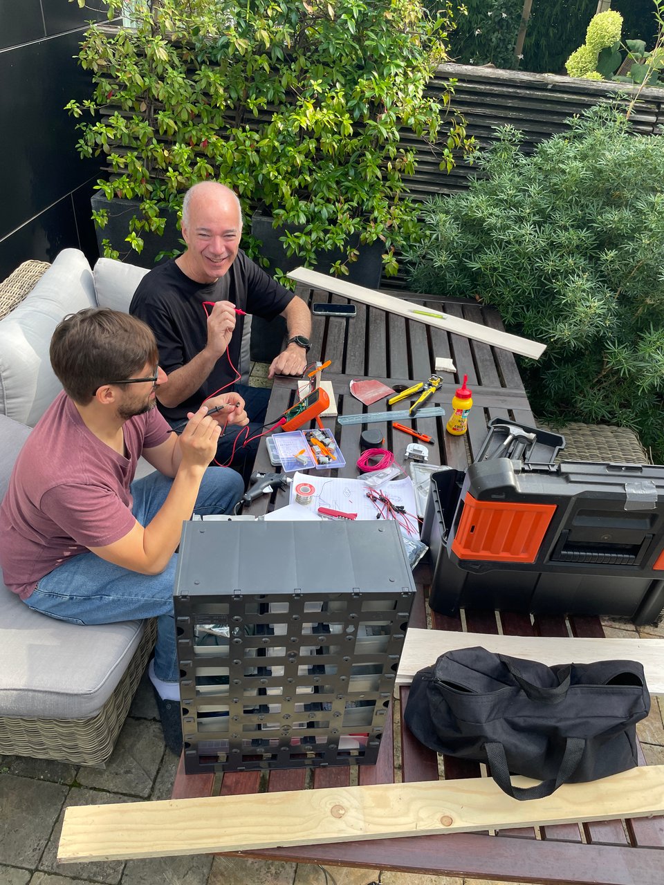 Two men sit at a table checking electrical connections on a project with a voltage meter.