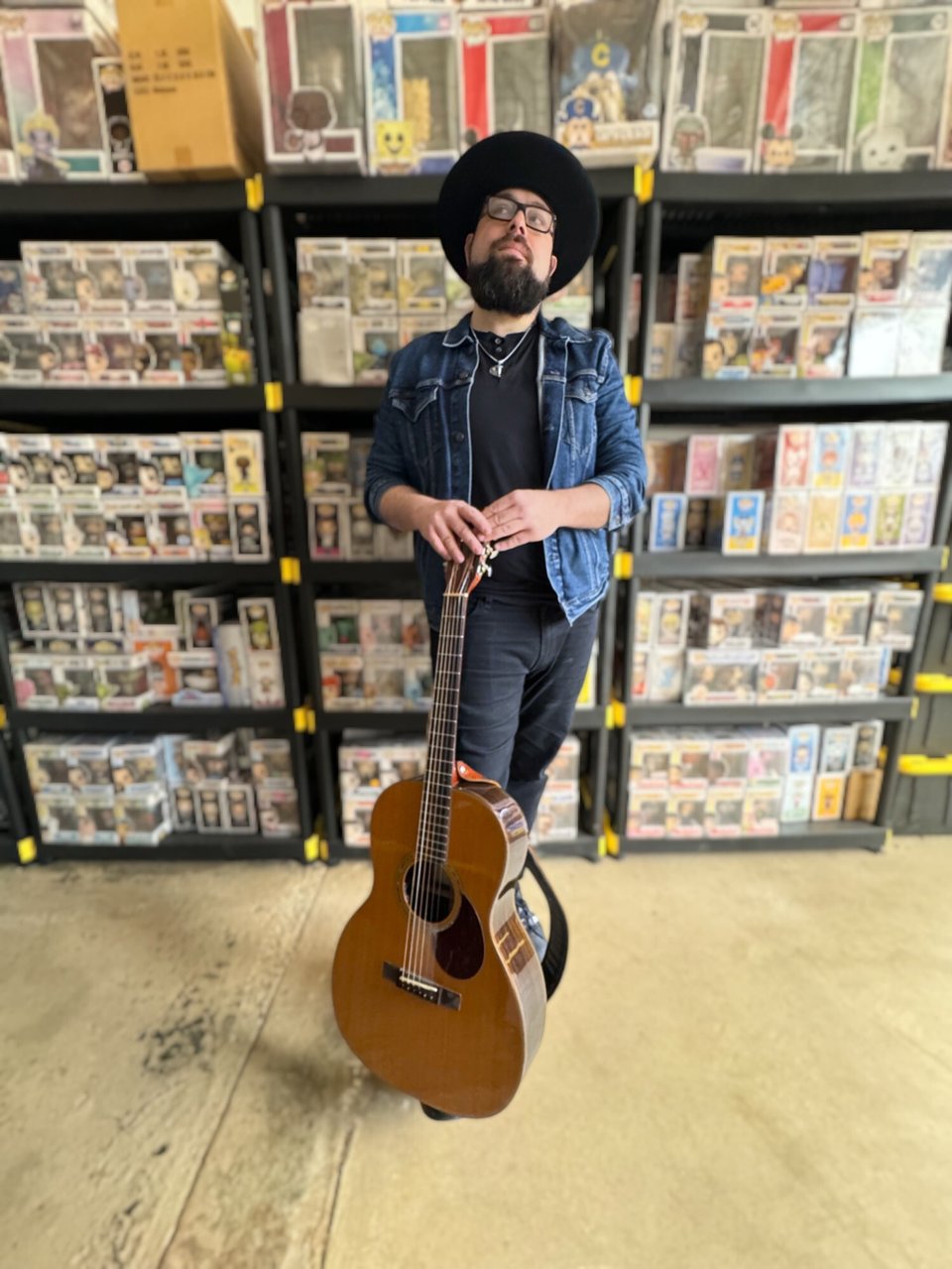 A person with a beard, wearing a broad-brimmed hat, a denim jacket and a black t-shirt, stands with his hand on the headstock of a vertical acoustic guitar.