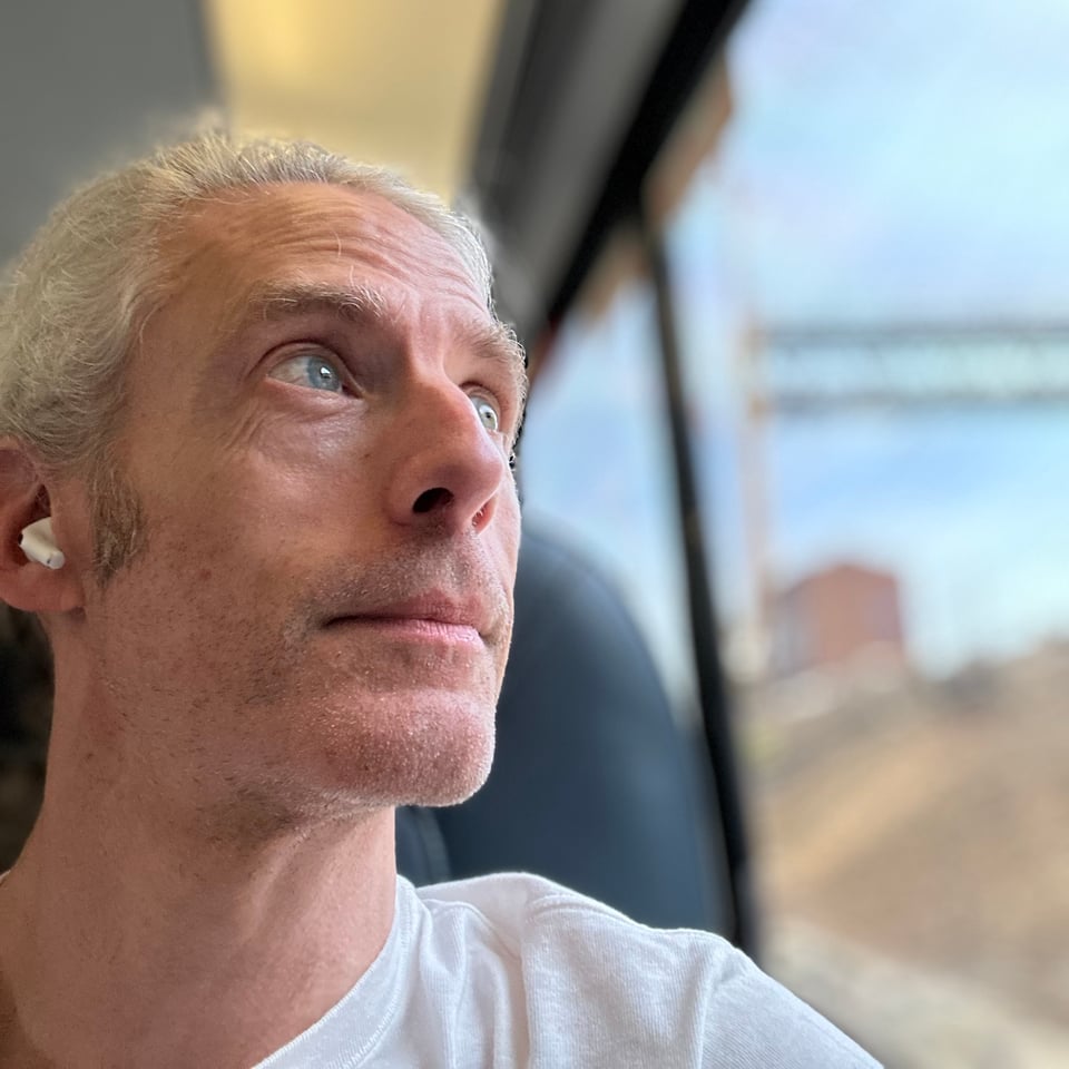 selfie of a white man with AirPods looking up & to the side, out the window of the train