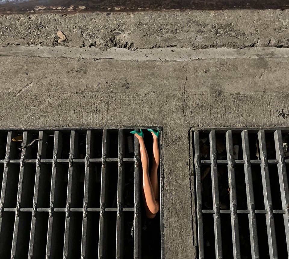 detached dolls legs wearing high heels stuck in the slats of a drainage grate in the sidewalk