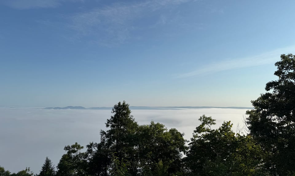 photo of fog-bound valley, taken from a mountain peak above the fog. Some hills/crests are slightly visible on the horizon, and the sky is hazy blue, but he fog below looks like sea mist, even though it's inland