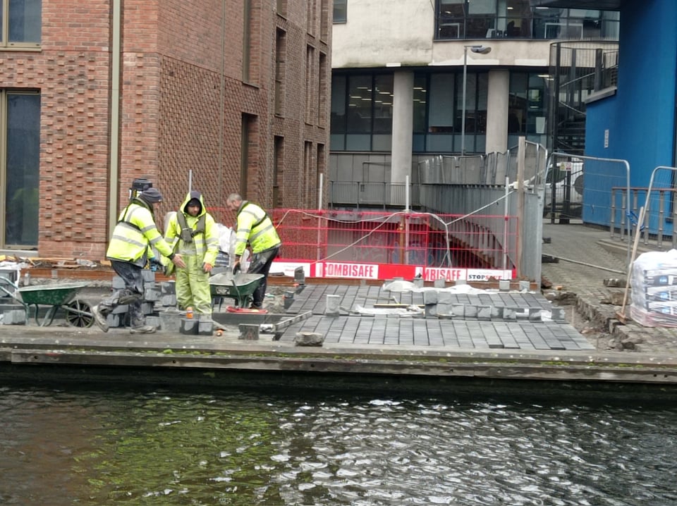 Three workmen in yellow high-vis jackets labour by the dark waters of a canal. They are in a fenced-off area laying grey flat-topped sett stones row by row.