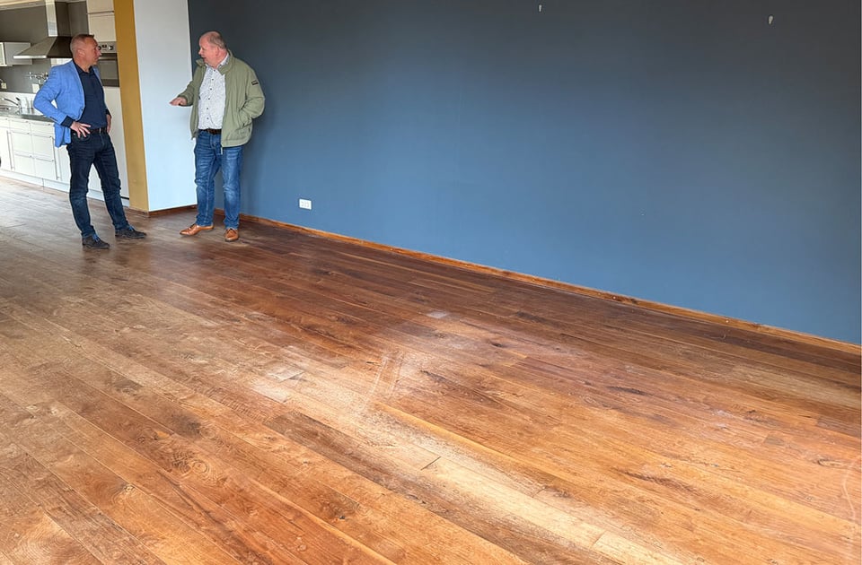 Two men chat in an empty room with a blue wall and bare wooden floors.