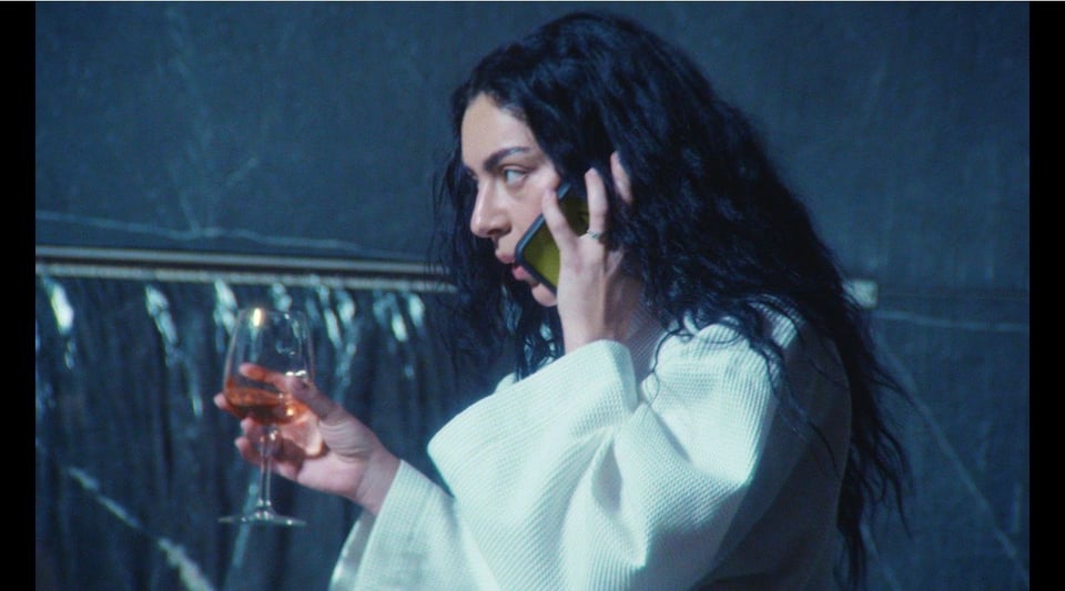 Charli in profile (long wavy dark hair draped over shoulders) wearing white bathrobe, glass of rose in left hand, holding iphone to ear, lips parted as though speaking.
