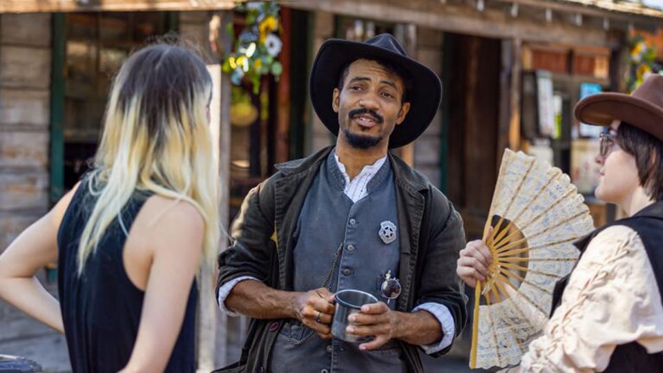 An old west sheriff wearing a black hat, carrying a tin cup, talks to two residents of an old west town.