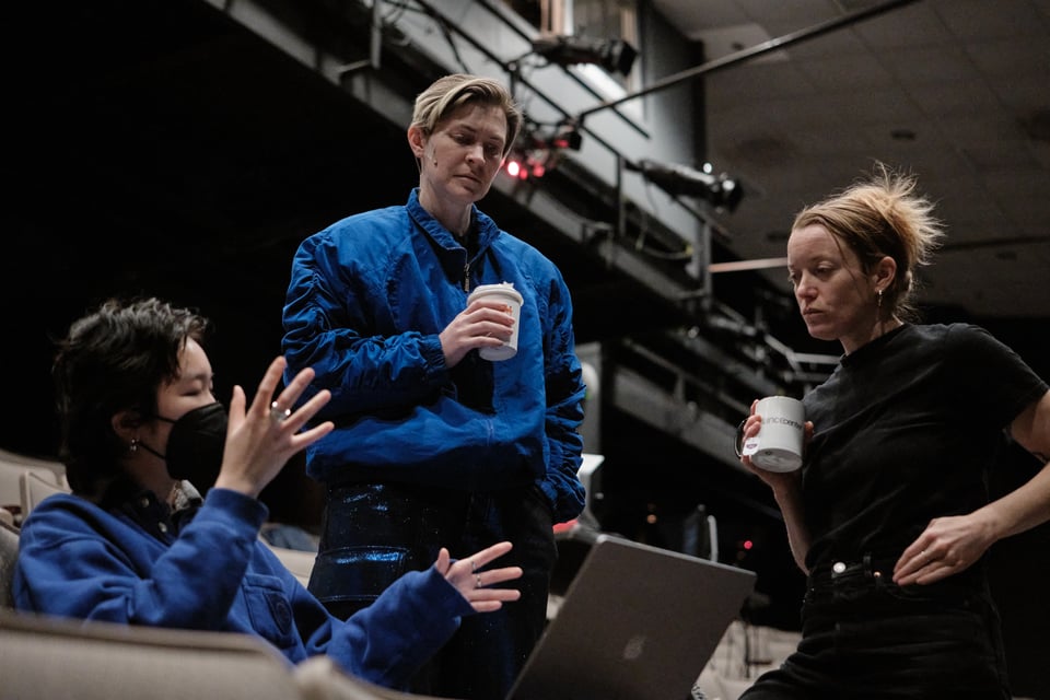 Ruby Que, Nora Sharp, and Jessi Barber stand or sit around a design they're looking at on a laptop. Ruby wears a mask and gesticulates while describing the design. Nora and Jessi each hold a coffee or tea and are listening pensively.