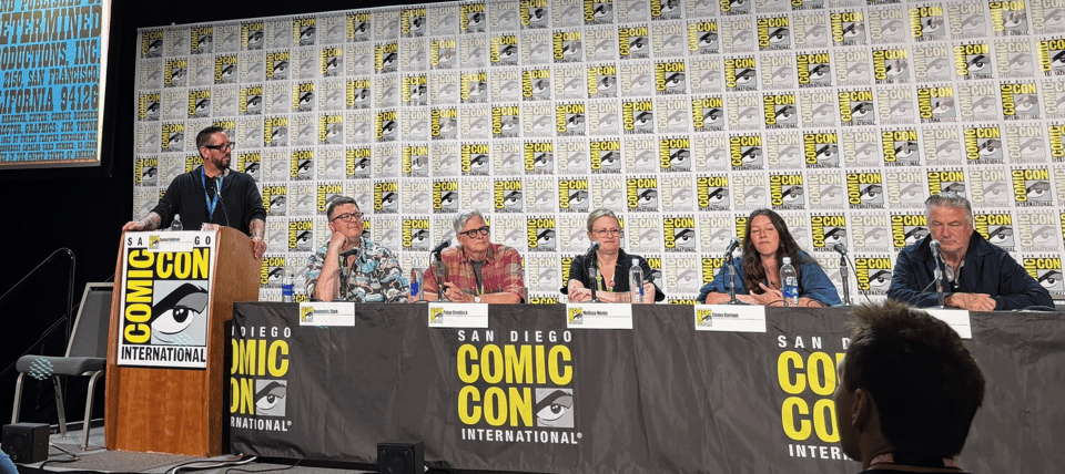 At the 2025 San Diego Comic Con, five people sit behind a table as a panel of experts, and a sixth person stands at a podium to moderate. Behind them all is the repeating pattern background with the logo for San Diego Comic Con.