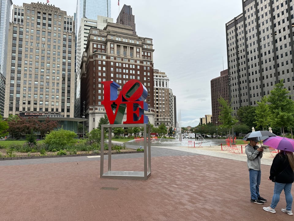 Photo of the LOVE sculpture in Love Park in Philadelphia, with two people standing nearby holding umbrellas
