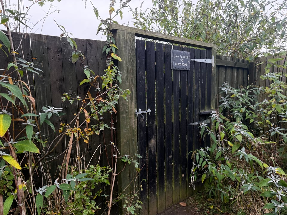 A black wooden slatted gate in a fence with a slate sign saying "Core path R138 to Flass Road"