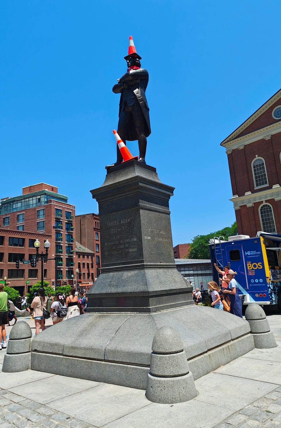 a photograph of a life-sized bronze statue of Samuel Adams standing atop a very tall plinth outside Faneuil Hall in Boston. Adams is posed contrapposto with his arms crossed across his chest. Somehow, someone has climbed up and put a bright orange traffic cone on his head, a red soccer scarf around his neck and another traffic cone askew between his feet. His dignity is unaffected by these shenanigans