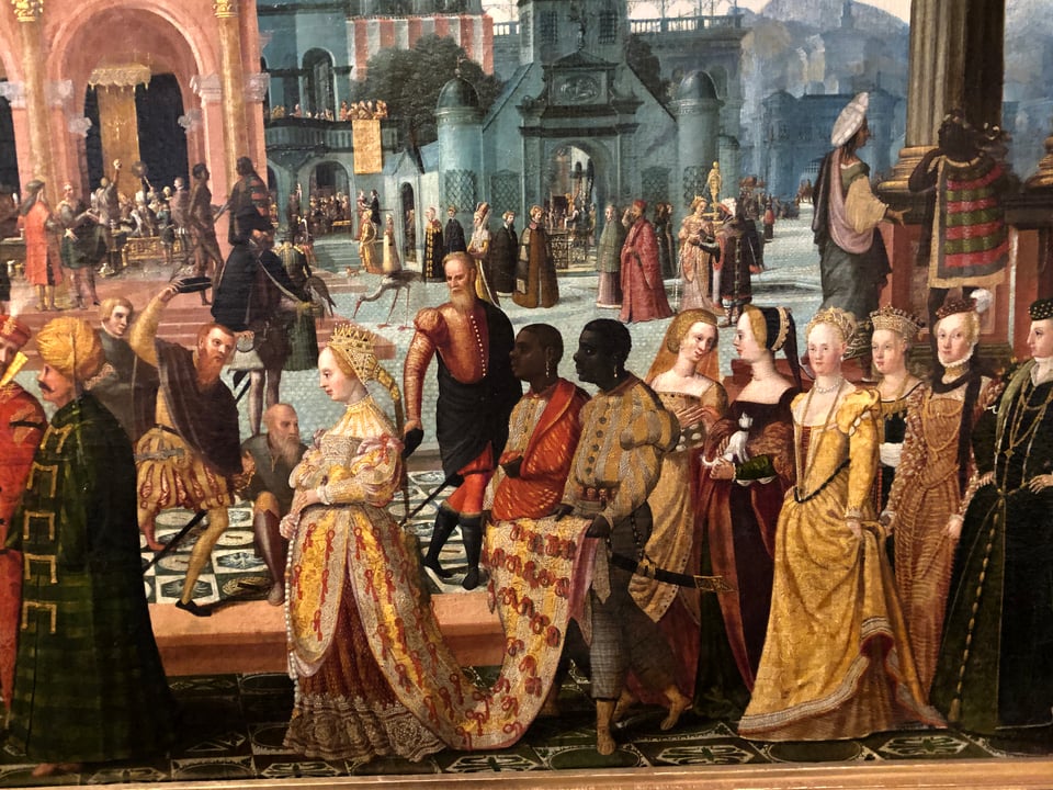 Richly dressed people in historic, vaguely Renaissance-style clothing process from right to left in the foreground. From left to right, they include men in turbans, a woman in a tiara and a dress with a long train helf up by a figure of African descent, equally richly dressed, who walks with another African figure. Behind them are an array of finely dressed women. Figures in the background include some darker-skinned figures; most of the figures in the painting are white. Two of the darker-skinned figures wear striped capes.