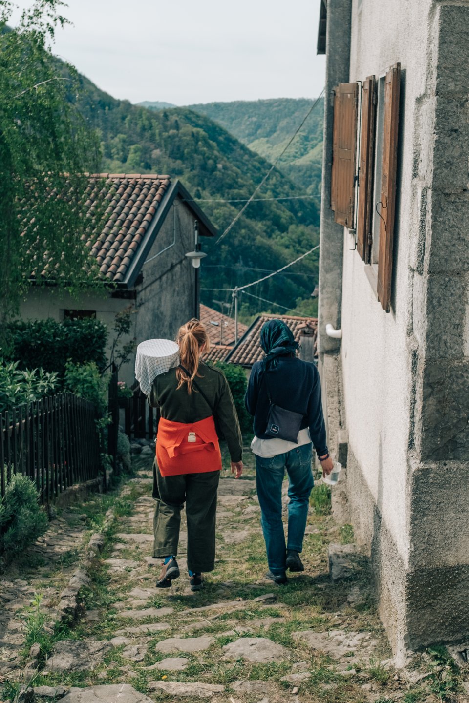 [Two friends walk down a stone path in the village. Concrete houses with terracotta roofs line the path. The mountains can be seen in the distance.]