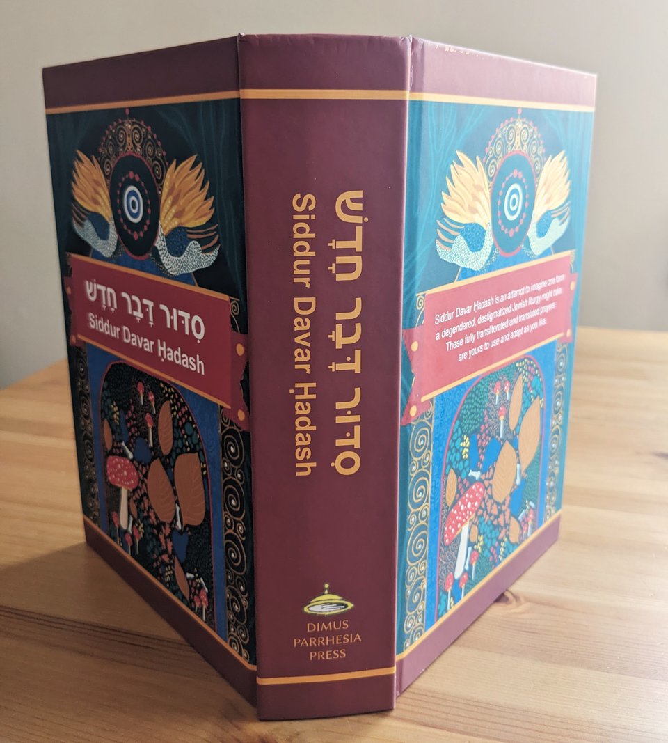 The siddur standing up on the table, spread so both the front and back covers are visible beside the spine. It's thick, solid, a really full volume.