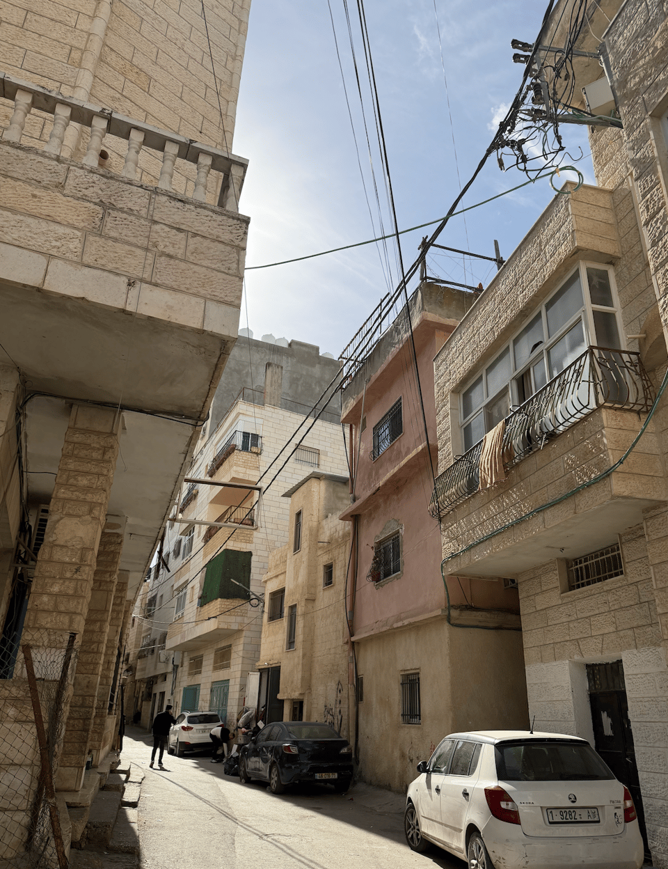 Photograph of a narrow urban street lined with multi-story residential buildings featuring stone and plaster facades in beige and light brown tones. Several parked cars and a few pedestrians are visible, with overhead utility wires crossing above the street under a partly cloudy sky.