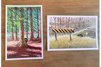 A photograph of two 4 x 6 mini prints. The prints are of paintings depicting small black blob creatures with white eyes. In the first painting, the creatures are in a park peeking out from behind two trees. In the second painting, two creatures are crawling out of the ditch at an abandoned rail crossing.