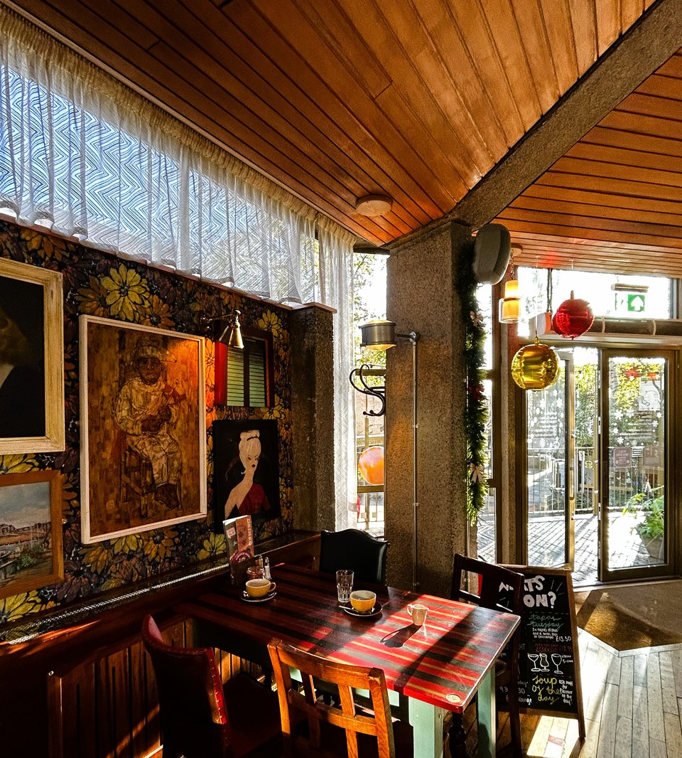 A corner of the interior, showing distressed 1970s floral wallpaper and an assortment of differently sized midcentury paintings. Thin curtains have been hug that have a white wavy pattern in them.
