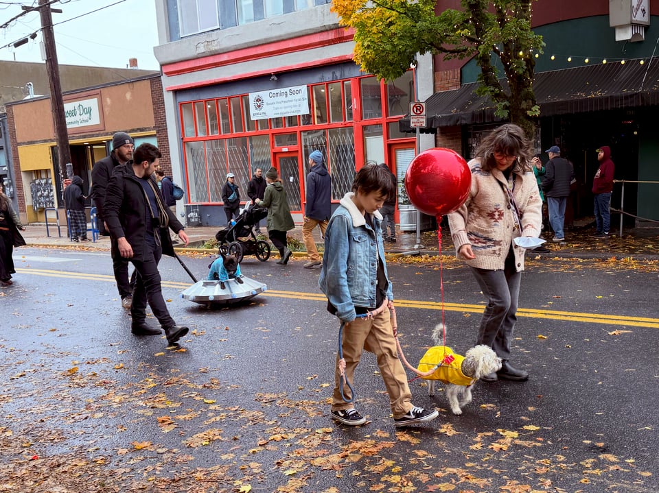 People walk dogs down a street. One dog is dressed as Richie from IT, wearing a rain coat with a red balloon tied to it. Behind that group, someone pushes a UFO cart on which a dog in an alien suit is riding.