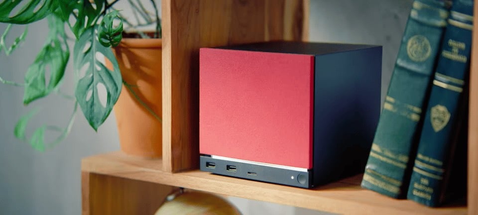 A compact computer with a red and black design is placed on a wooden shelf next to a potted plant and old books.