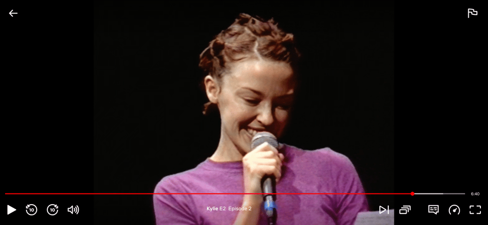 Screenshot from a TV programme. We see Kylie Minogue against a black background. She's holding a microphone and smiling.