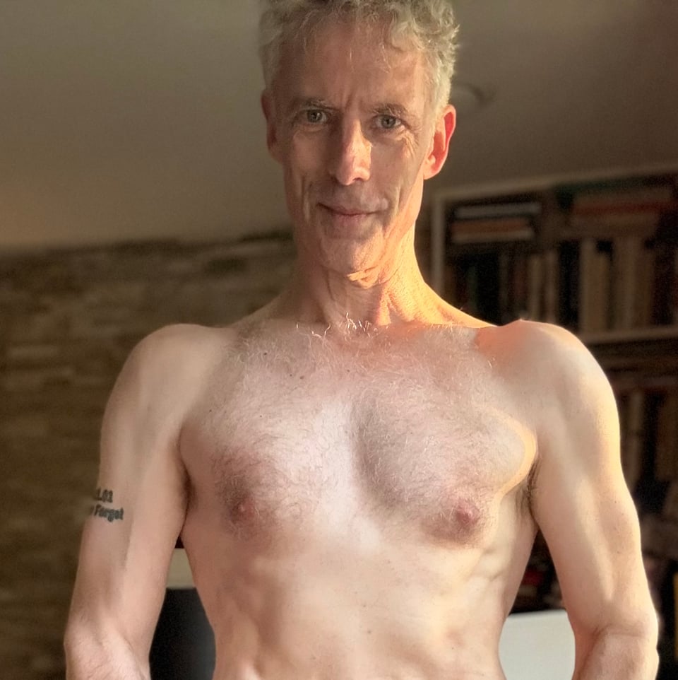 Portrait-mode selfie of shirtless white middle-aged man from sternum up. He's smiling, and there's sunlight on the left side of his neck and face, and his muscles look pretty swole for his build & age