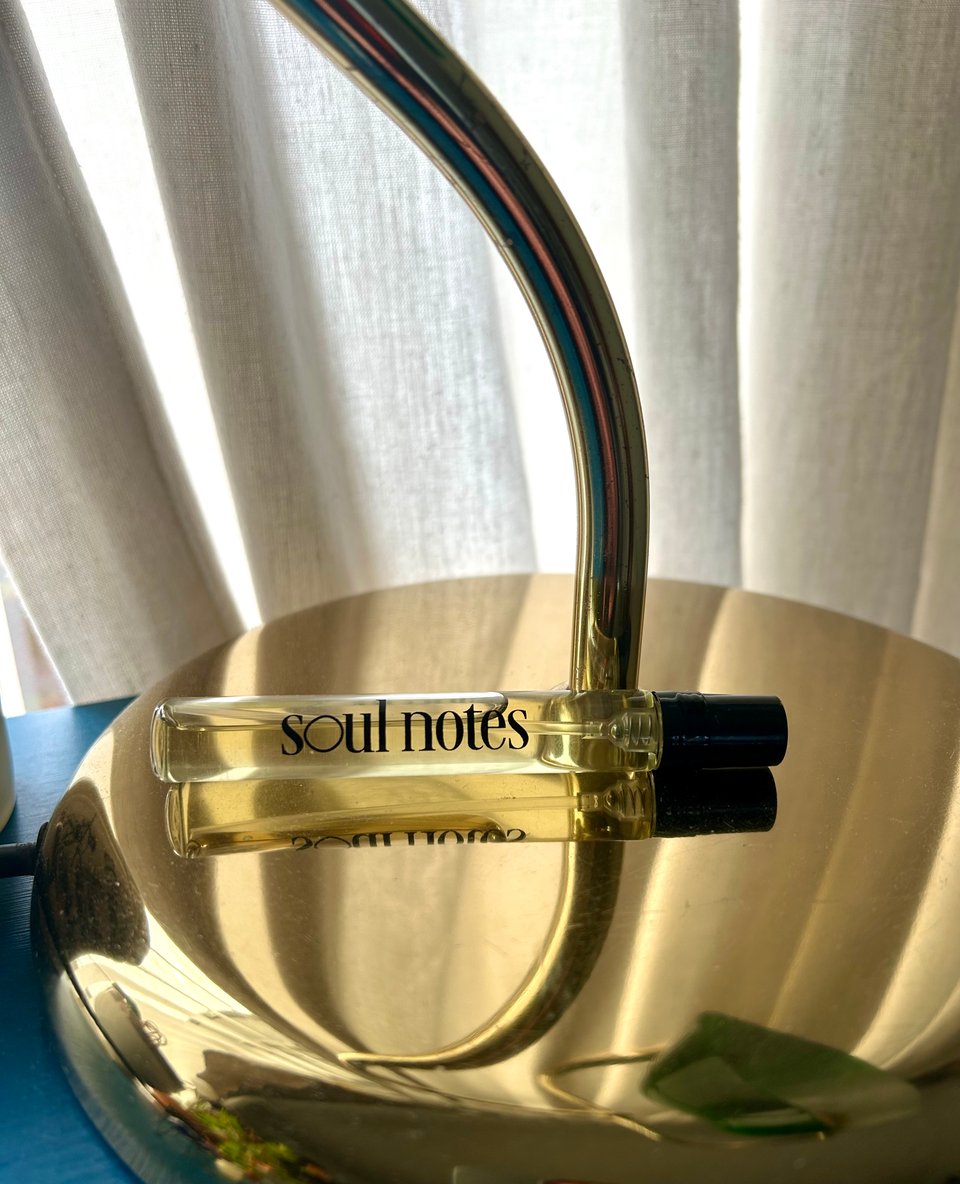 A glass travel size bottle of Soul Notes perfume is on a brass lamp base. Behind are cream cotton curtains. Image by Rowan Ambrose.