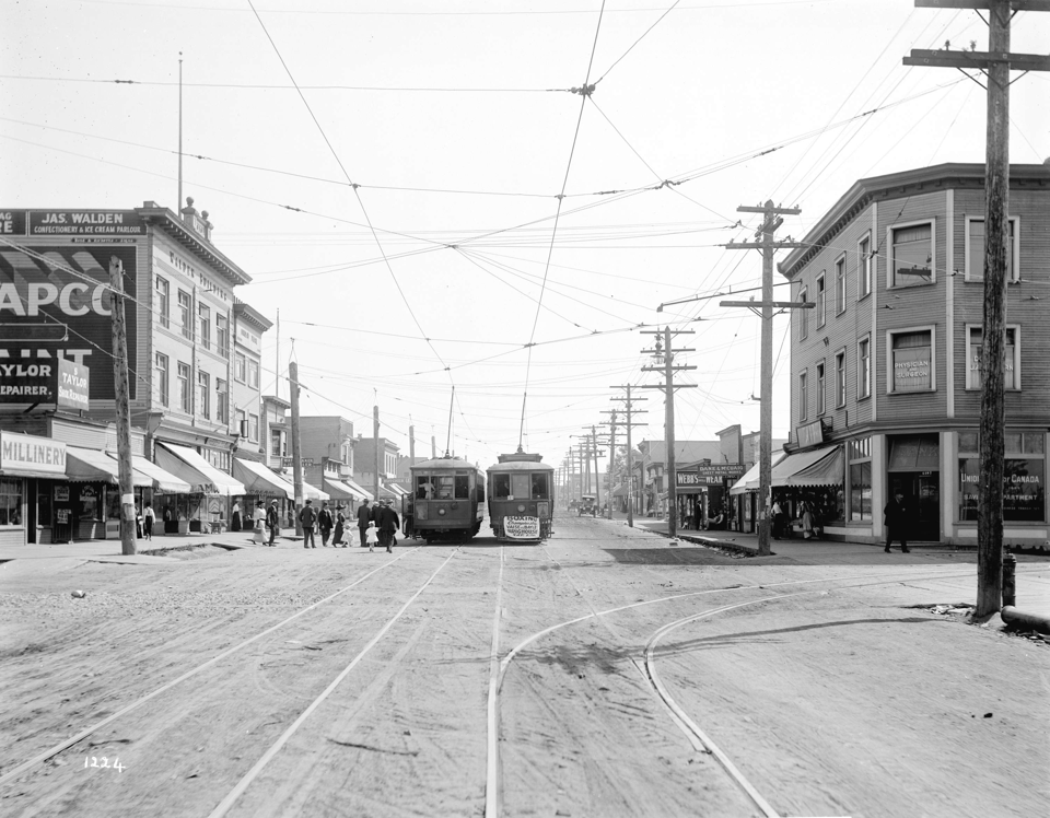 View of a wide dirt road with street car tracks. Buildings alongside and, in the centre, two streetcars are parked next to each other with people trying to board.