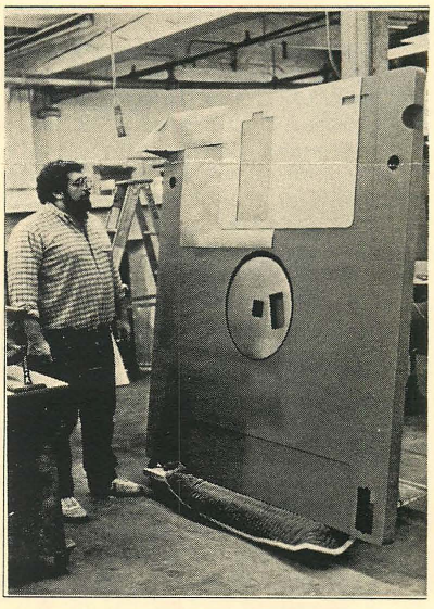 A bearded man in glasses, a buttonup shirt, and jeans is standing next to a 6-foot tall model of a 3.5" floppy disk in a warehouse.