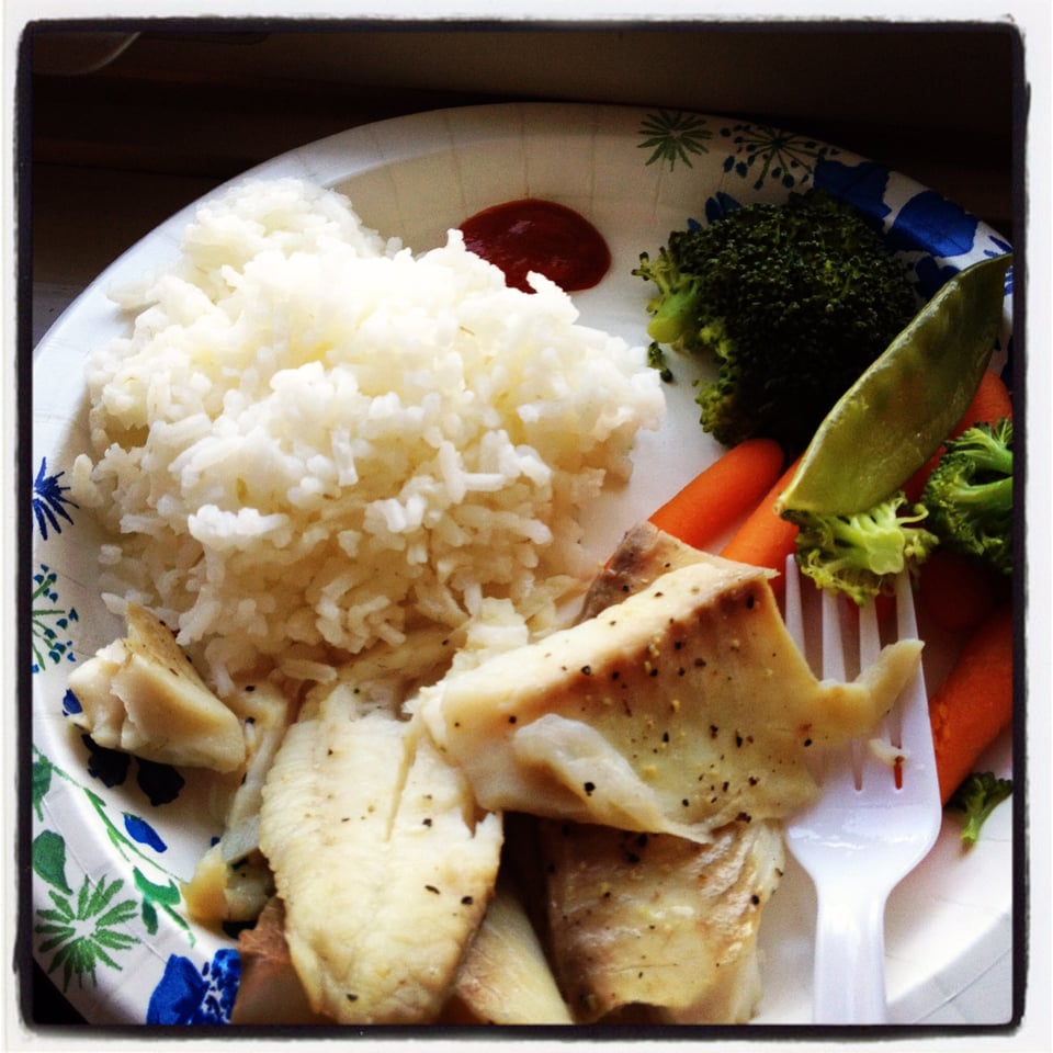 A paper plate loaded with poorly cooked rice, steamed vegetables, and a lightly seasoned tilapia fillet.