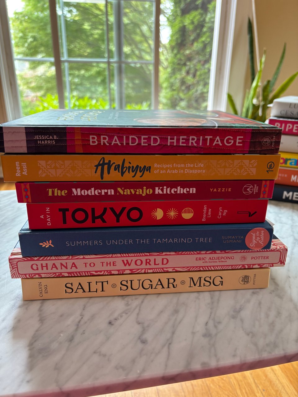 A stack of books the author purchased in one BL visit, including copies of Braided Heritage, Arabiyya, The Modern Navajo Kitchen, Tokyo, Summers Under the Tamarind Tree, Ghana to the World, Salt.Sugar.MSG.