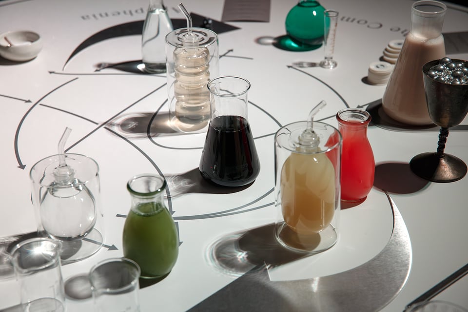 A series of beakers and bottles full of different coloured liquids, pink and green and almost black. None of them are labelled. On the table is a printed diagram with mysterious arrows, pointing to different parts of the table.