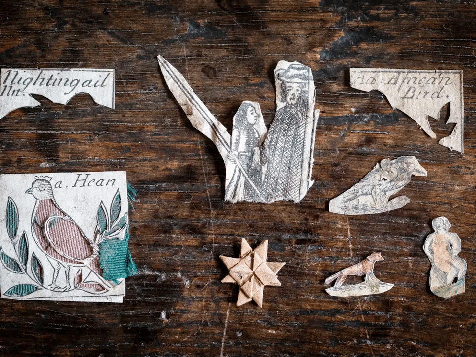 A photo of several paperwork artifacts. One is an origami eight-pointed star; the others are cutouts of images such as as bird, a person with a long pole, and a small animal. They all look very old