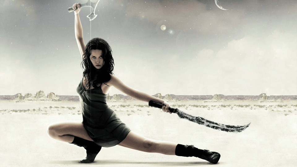 Summer Glau, a white girl wearing a short dress, does a pose with one leg outstretched and the other one bent, holding a sword in the air and another one parallel to her leg. She's in a desert landscape