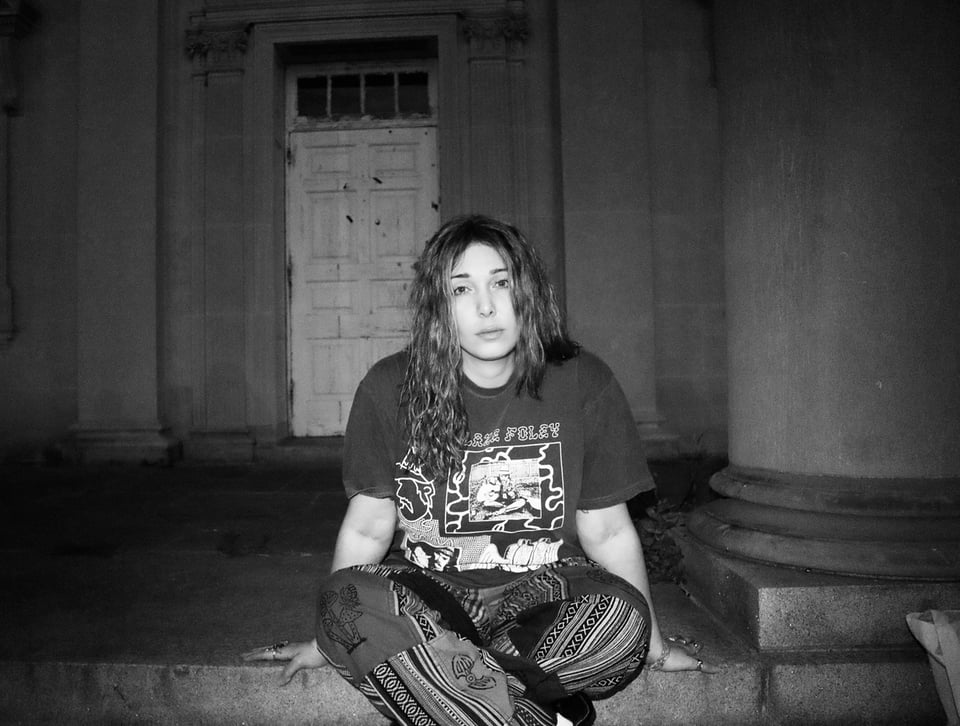 A black-and-white photo of a woman with long, brown hair sitting on a step in front of a door. She's wearing a t-shirt and motley pants.