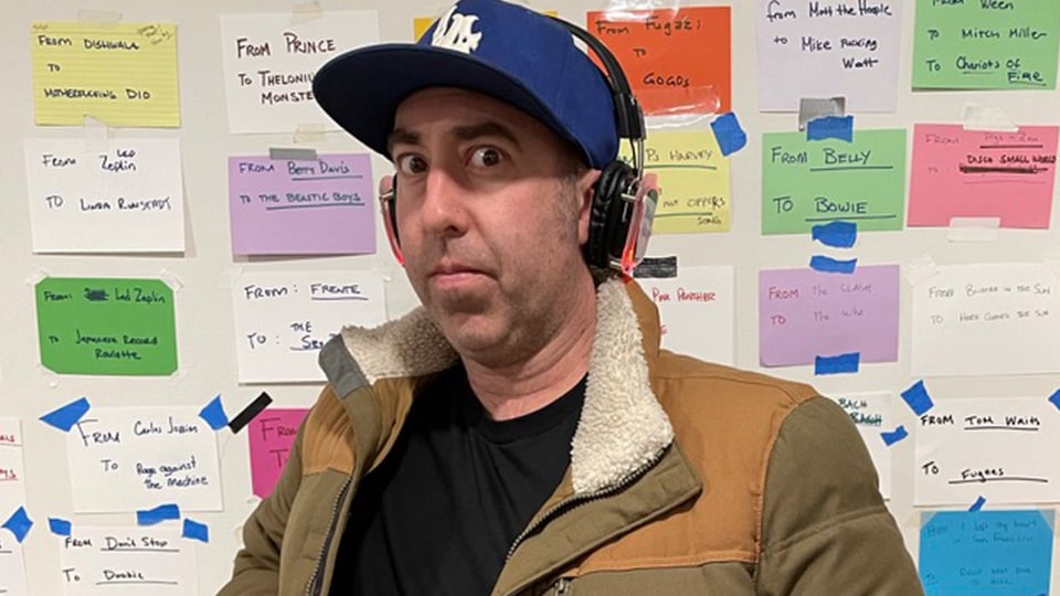 A man in a blue dodgers cap and headphones and a sheerling jacket, stands in front of a wall of colored index cards.