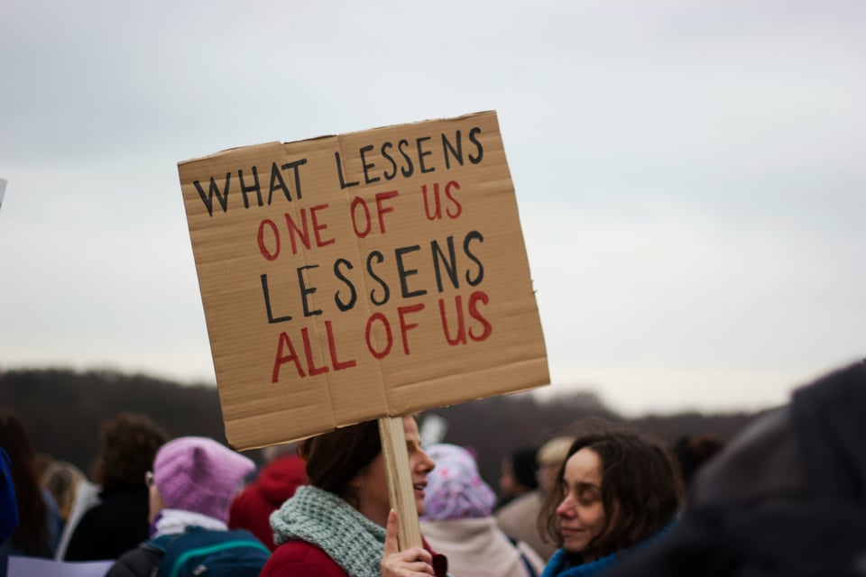 Cardboard protest sign: "What lessens one of us lessens all of us."
"What lessens" and "lessens" are written in black, "one of us," "all of us" are written in red.