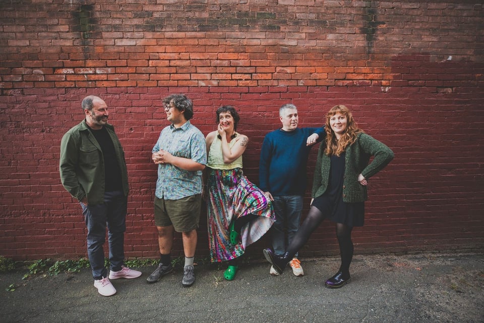 Three men and two women stand in front of a brick wall. Each woman has one foot off the ground, as if they're comparing footwear.