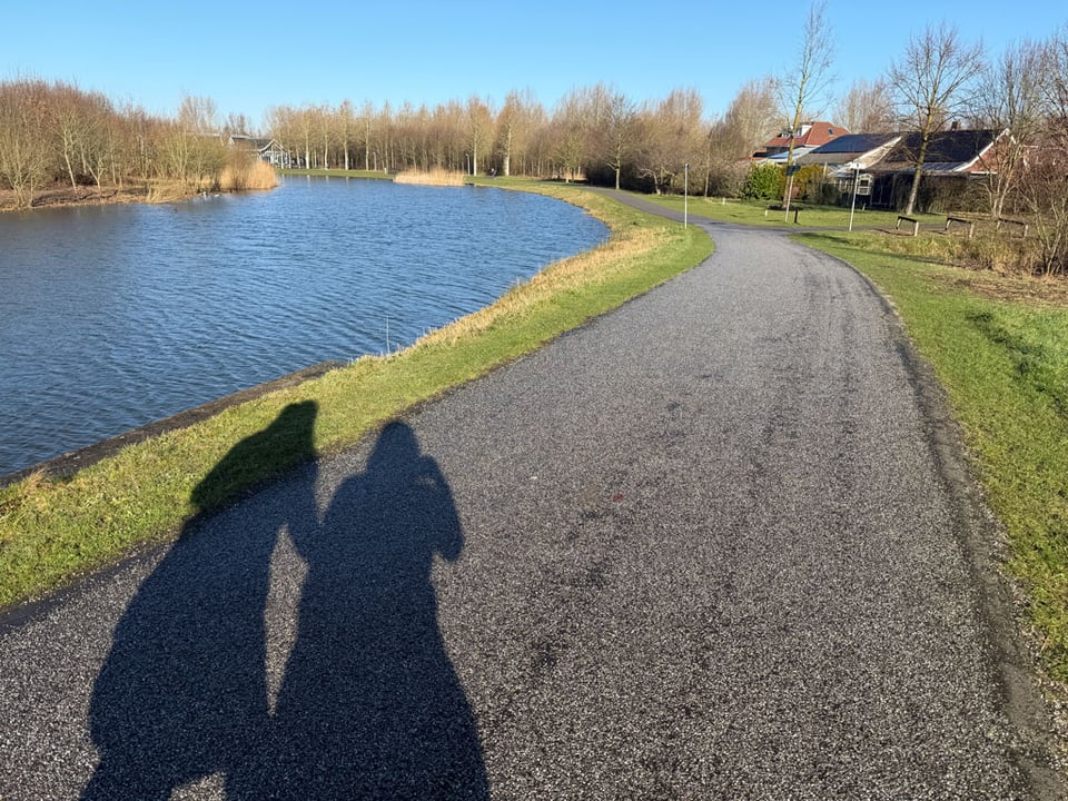 Two shadows of people walking along an asphalt footpath in a park next to a canal of water.