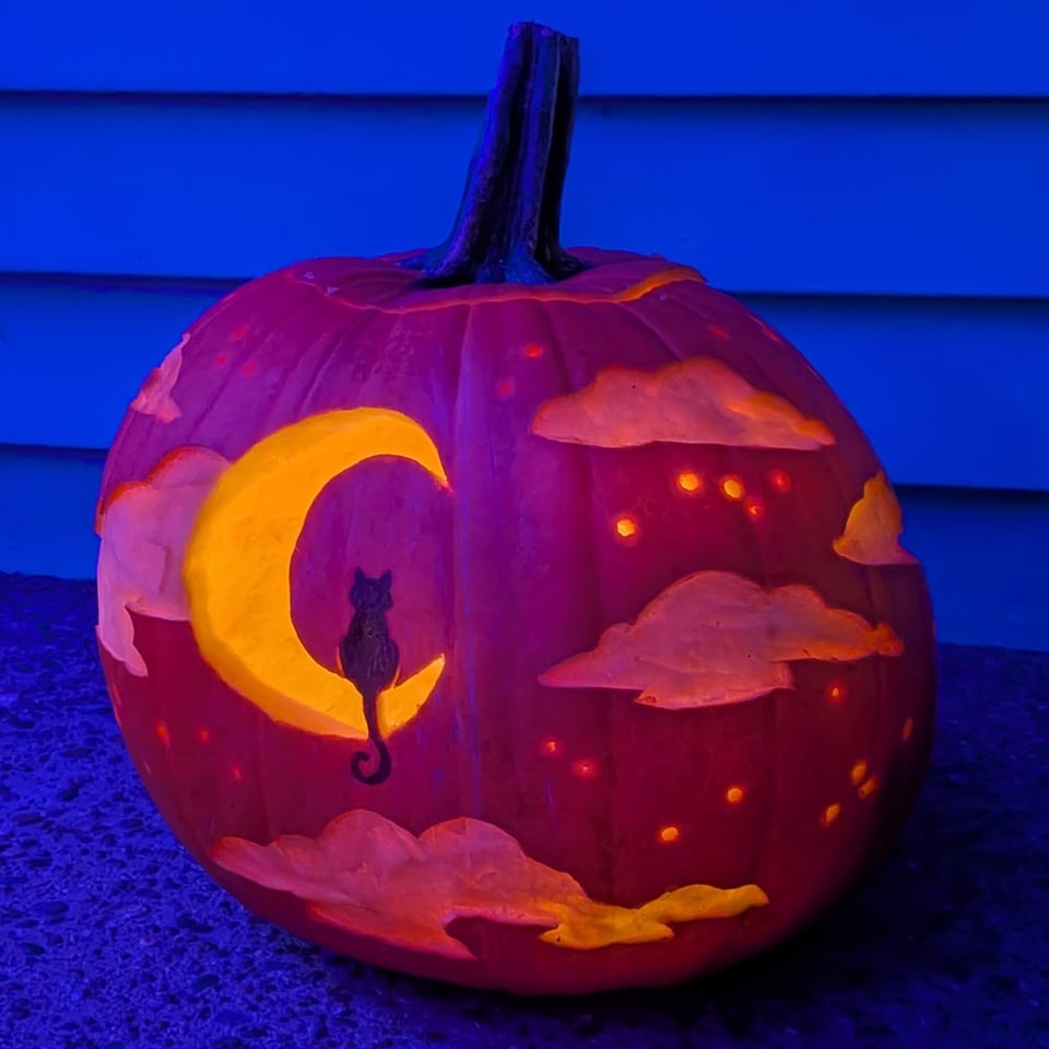 a carved pumpkin depicting a black cat sitting in silhouette on a crescent moon surrounded by fluffy clouds and a scattering of stars.