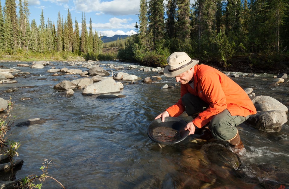 A man wearing a beige hat, orange jacket, and rubber boots kneels in a shallow, rocky river surrounded by evergreen trees and distant mountains. He is panning for gold, holding a black pan filled with water and sediment as he carefully swirls it. It's a pretty serene image