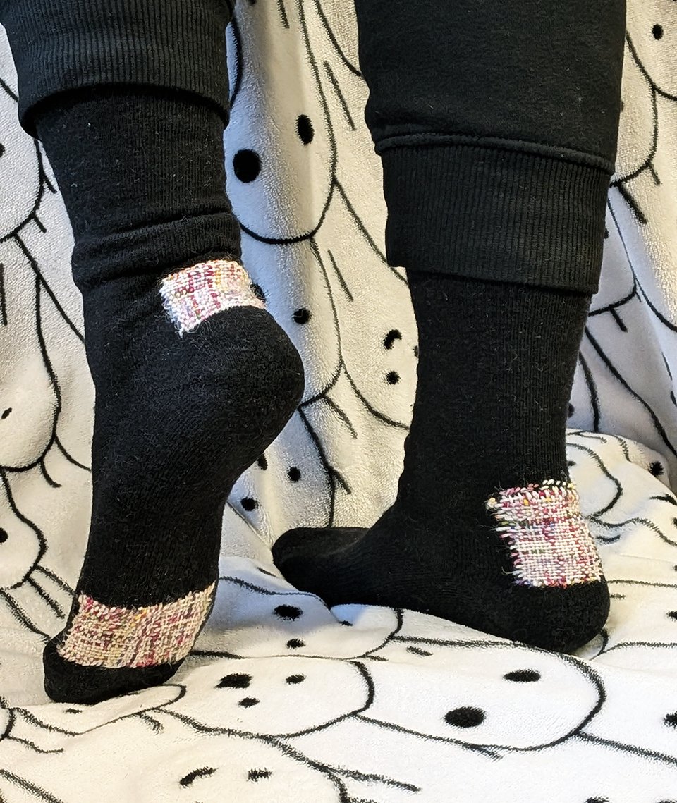 Maia's feet are swathed in black wool socks with large, neat darned patches on the heels and ball of the foot. The darned patches are white with speckles of other colors. The backdrop is a blanket with ghost faces all over it.