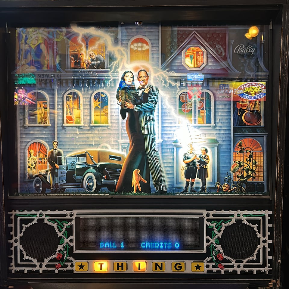 The backboard of an Addams Family pinball machine with Gomez and Morticia hugging in front of the mansion while other characters look on.