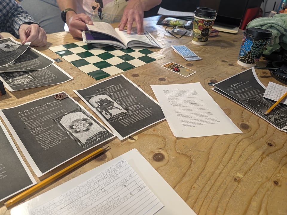 An overhead view of a tabletop roleplaying game in progress, with several players gathered around a large, well-worn wooden table. Two players are visible from the shoulders down, one leafing through an open rulebook while another holds a printed game sheet.
The table is scattered with black-and-white character sheets featuring illustrations and descriptions of Tarot cards, including The Wheel of Fortune, The Emperor and The Star. A single colourful Tarot card lies face up beside a blue-backed deck of playing cards. A green-and-cream chequered board sits in the middle of the table.
Handwritten notes, pencils, two brightly decorated takeaway coffee cups and a pair of glasses add to the scene. The cluttered tabletop and players consulting their materials capture the atmosphere of a collaborative, face-to-face roleplaying session, with a distinctly occult or Tarot-inspired theme.