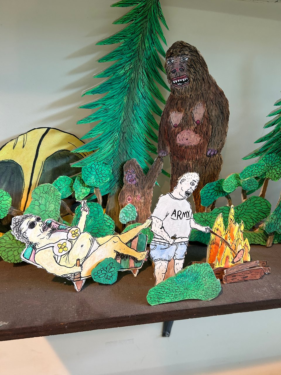 A paper diorama done in ink and colored pencil (I think). It depicts a large white woman in sunglasses and a daisy bikini, reclining on a chair with a beer and cigarette. A bald white man in jorts and an Army t-shirt roasts a hotdog over a fire. Behind them, a very large Sasquatch with drooping breasts holds hand with a child-sized sasquatch, both of them watching the humans with a vaguely longing expression.