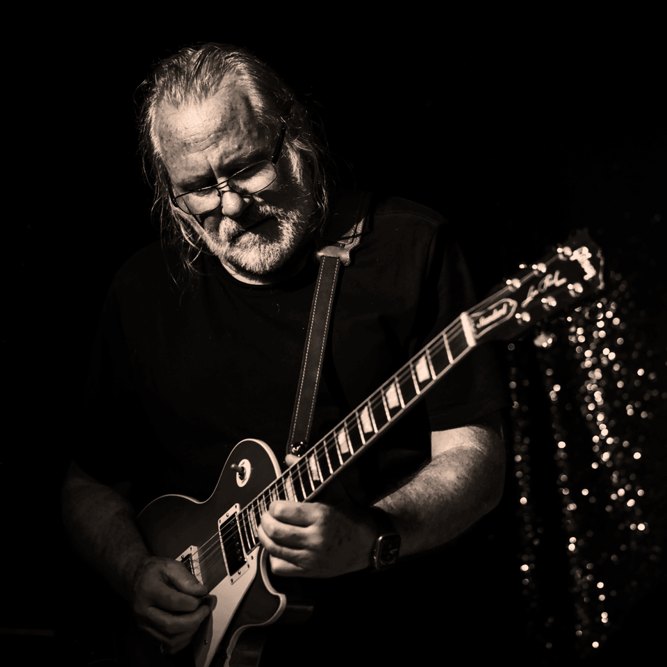 A man with thinning white hair, a beard and glasses plays a Les Paul guitar that looks as if sparks are cascading down off the headstock. The photo is sepia-toned.