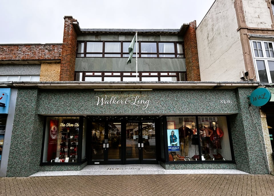 A shop front. The upper storeys are set back from the street line and appear to be a metal frame with ribbon windows and marble panels. The ground floor front is covered in blue, grey and green mosaic tiles.