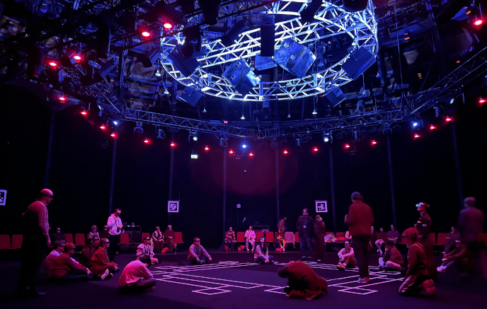 The set-up for Kagami at The Roundhouse, a London venue. Black curtains encircle the space; audience members are seated in red chairs encircling the space, and are sitting and standing in front of an area in the centre of the room marked off with white tape. The audience members are all wearing Magic Leap Twos. A large lighting rig hangs overhead. The room is lit in pink and blue lights.