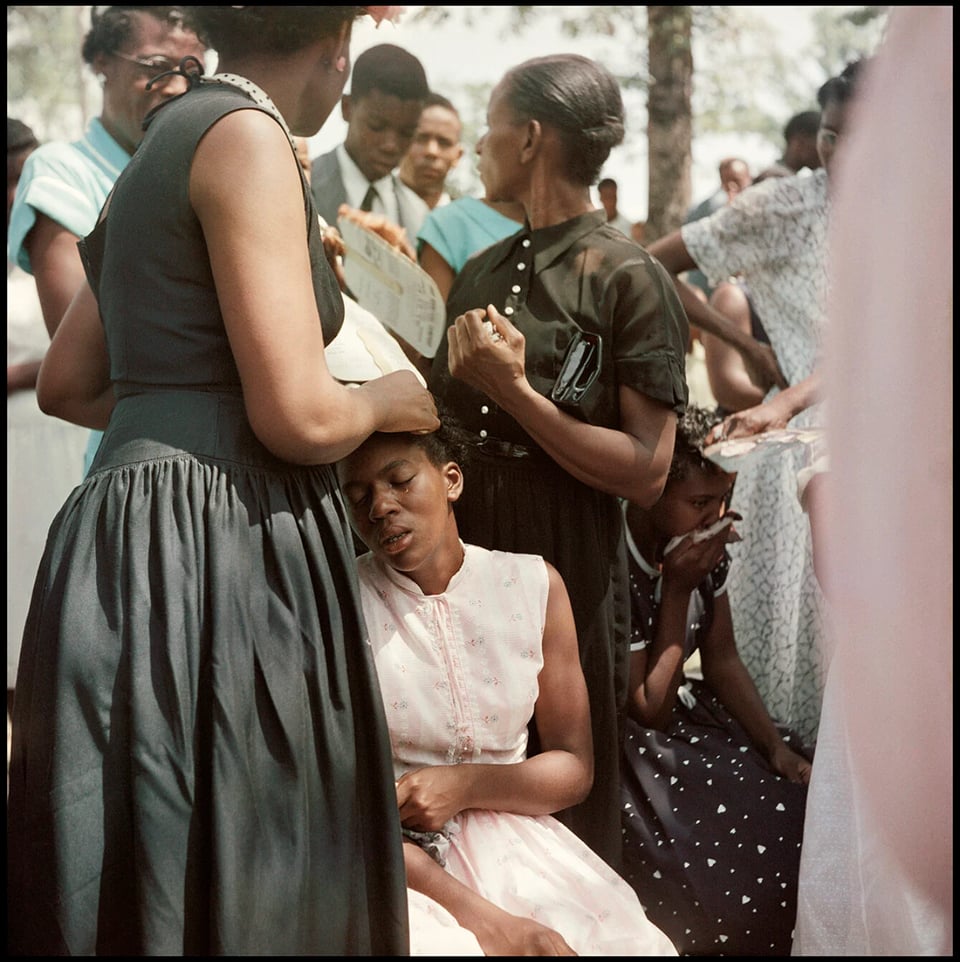 A photo taken by Gordon Parks of people at a funeral, with one young woman in the foreground with tears coming down her face as she is apparently comforted by another woman.